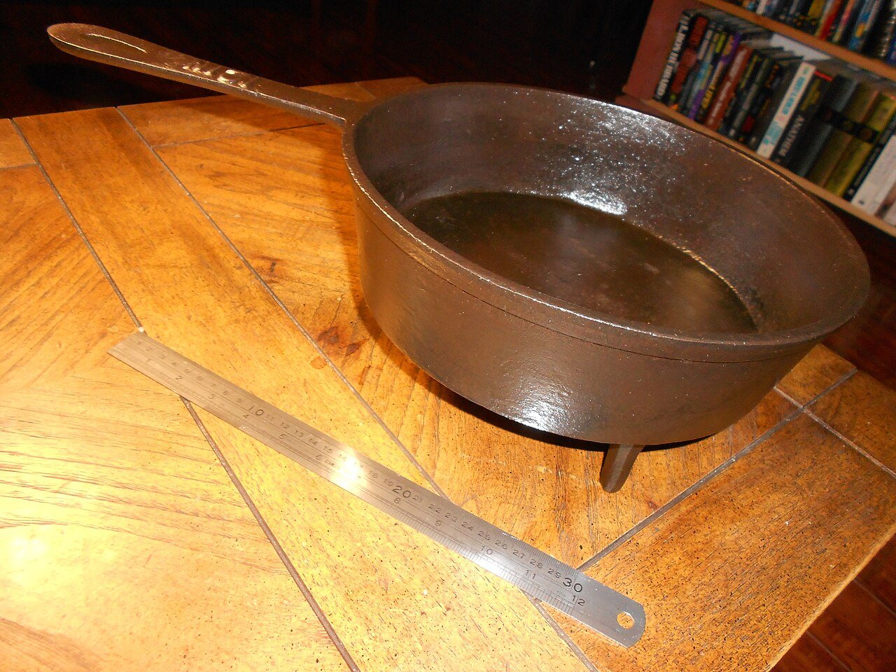 A 19th-century three-legged cast-iron skillet, known as a spider, on a wooden table next to a metal ruler