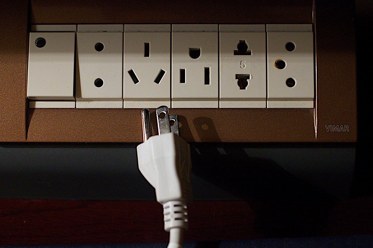 A white three-prong plug pulled partway out below a row of wall sockets for different plug types