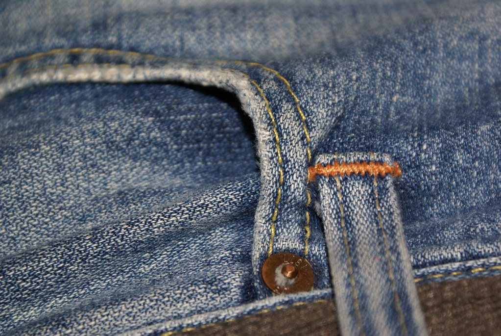Close-up of the front pocket of faded blue jeans, showing yellow stitching, a copper rivet at the pocket corner and a belt loop